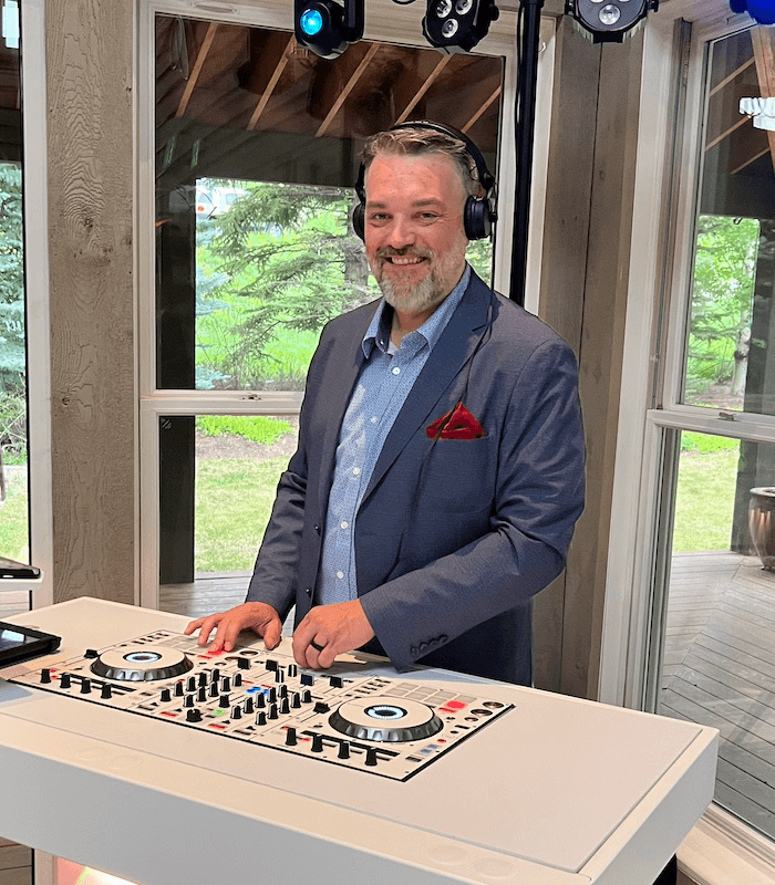 MATTHEW KAYS A man in a blue suit jacket with a red pocket square is standing at a DJ booth with headphones on, smiling. He is indoors with a view of greenery outside the windows.