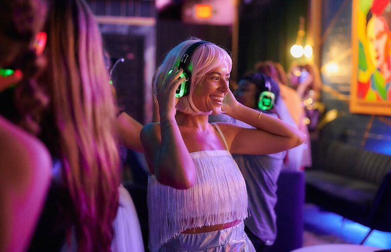 after party silent disco A person with a blonde wig and fringed top enjoys a silent disco, wearing light-up headphones in a dimly lit room with colorful lighting and background artwork.