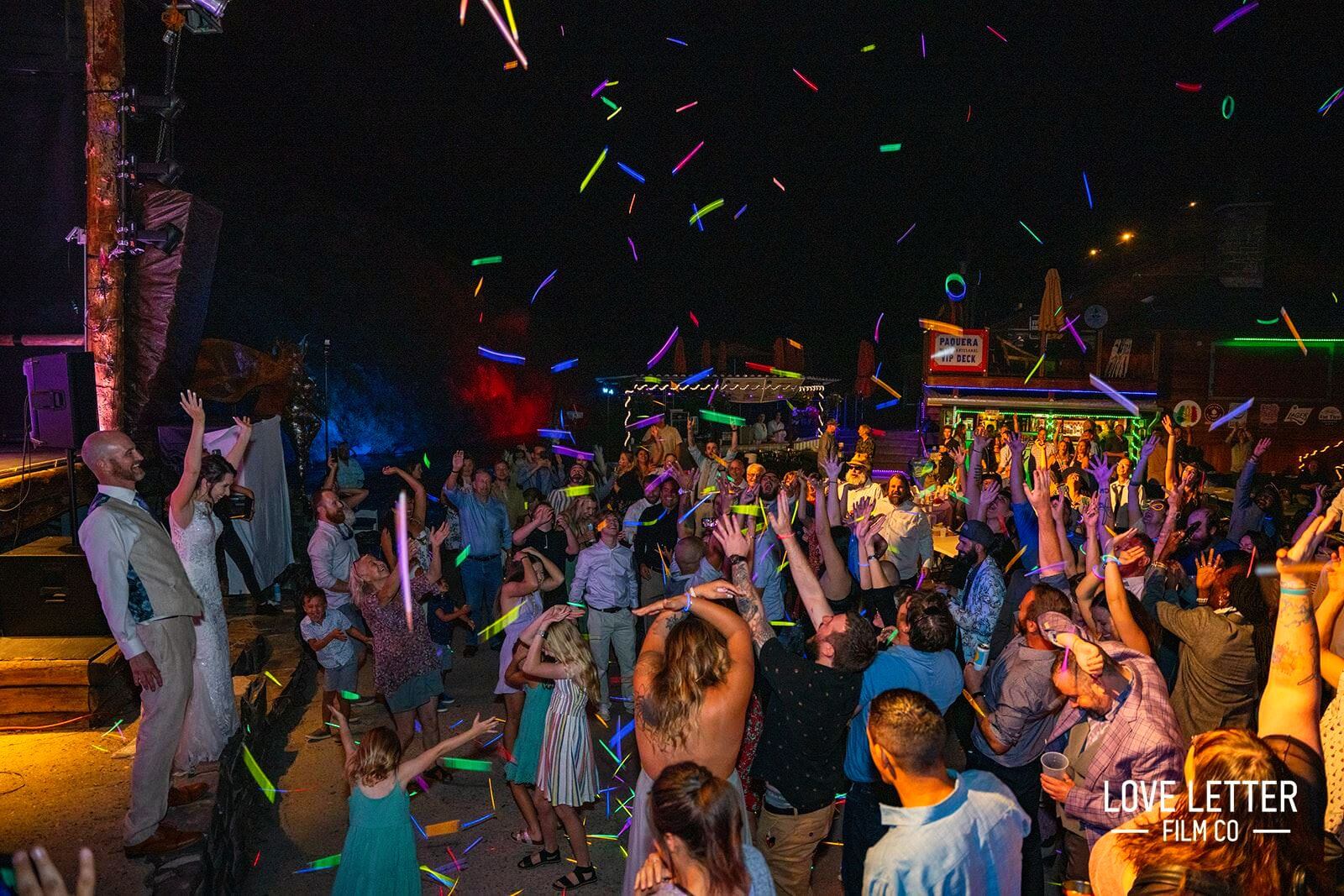 c69ec161-32f7-6ad1-fd77-3cf09e44d894 A large group of people dancing energetically at an outdoor night event with colorful lights and glow sticks.