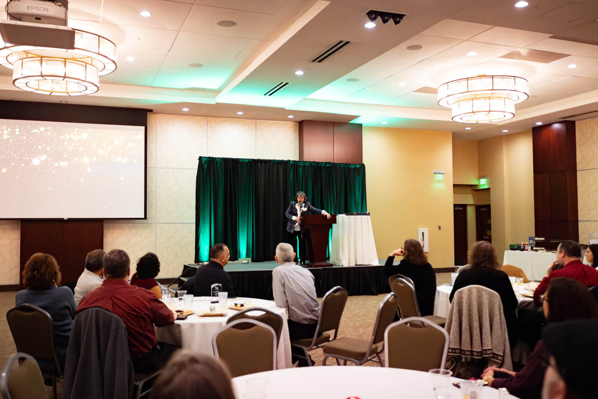 A speaker stands at a podium on stage addressing an audience seated at round tables in a banquet hall with green accent lighting.